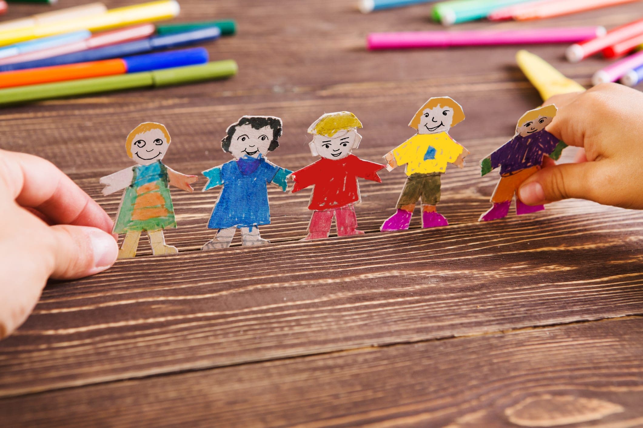 Hands holding a chain of hand-colored paper dolls on a wooden table strewn with markers
